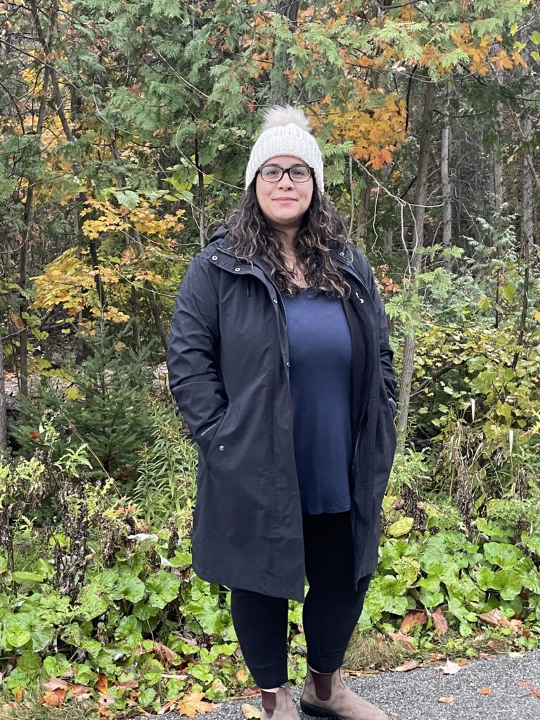 Jennifer Koutoulas, standing in front of some trees and other foliage. She is wearing a white hat and black coat