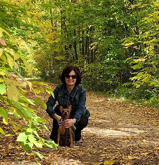 Kim Sexsmith crouches down to pose with her small dog while in the woods