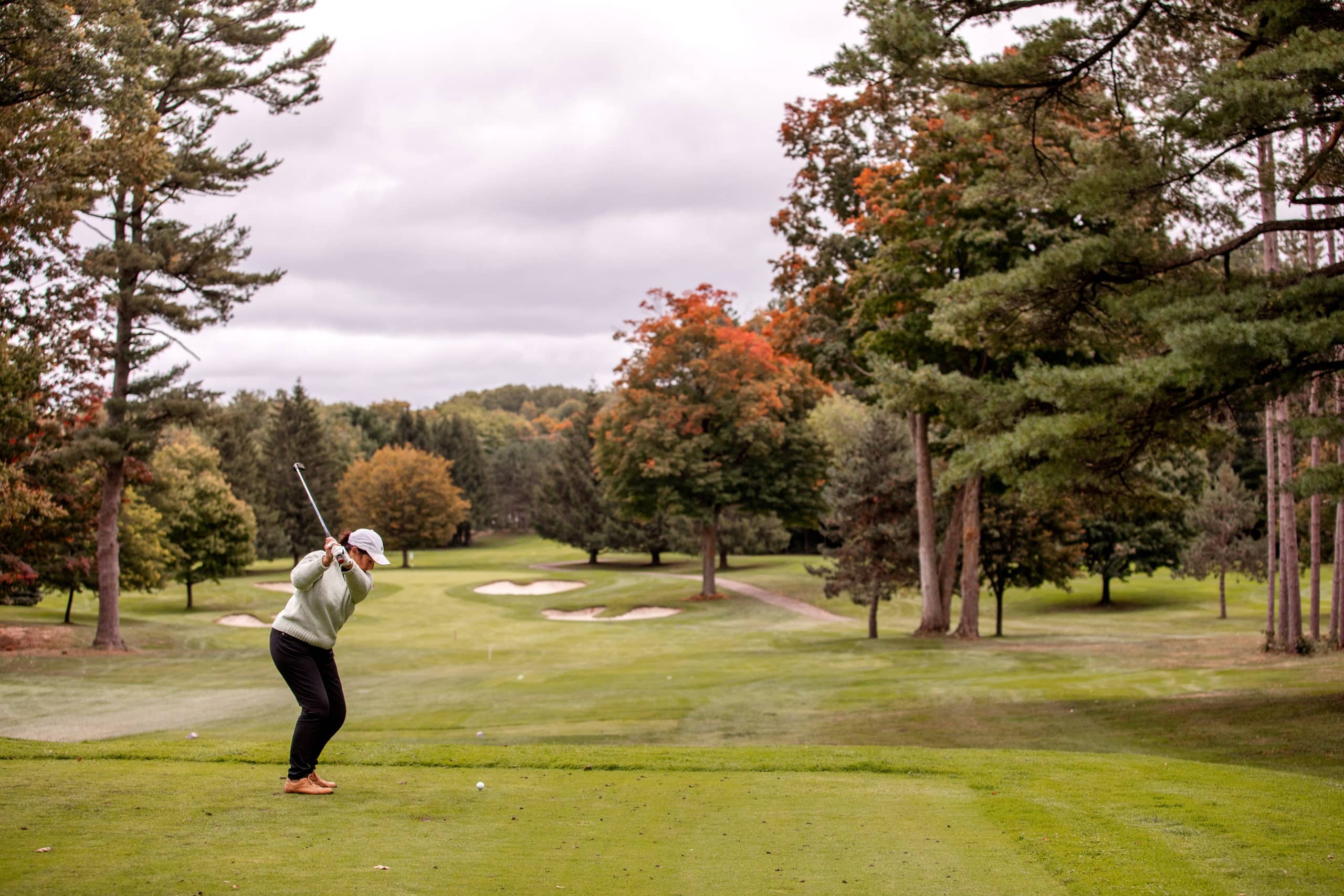 Lois swings her golf club in preparation to hit the ball of the tee block. The trees in the background are starting to change colour for fall.