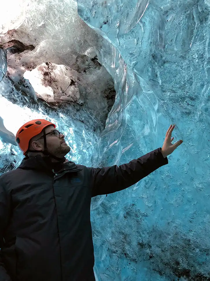 Ryan McLeod, touching a wall of ice inside an ice cave. He is wearing an orange helmet and black coat