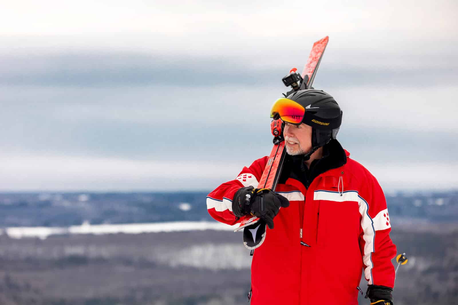 A man stands at the top of a ski hill in winter gear with ski's over his shoulder.